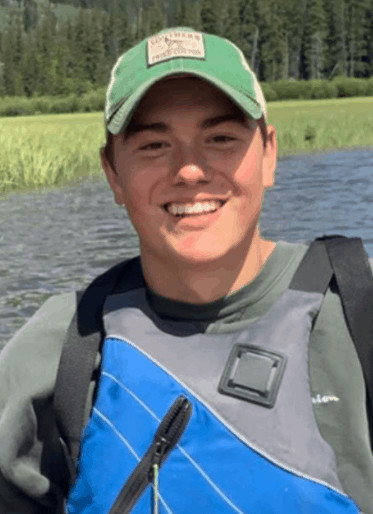 Tanner, a Makerspace staff, presumably on a boat. He has a green hat on and blue and grey life jacket.