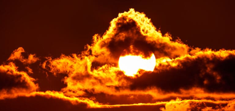 The bright sun, shining orange, with some clouds surrounding it.