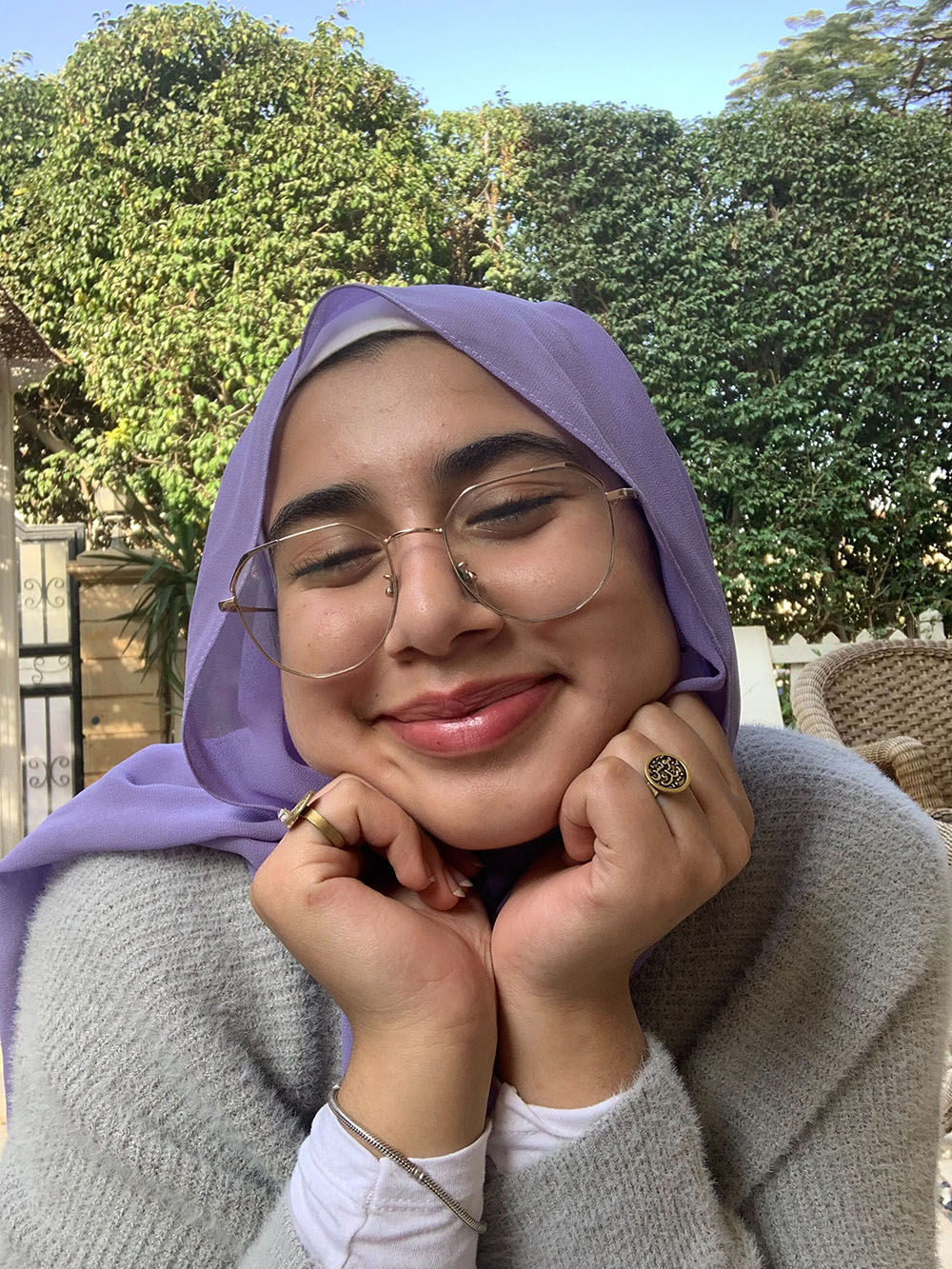 A portait of Somaya Salama, Her head is reasting on her hands with a smile on her face, with trees behind her.